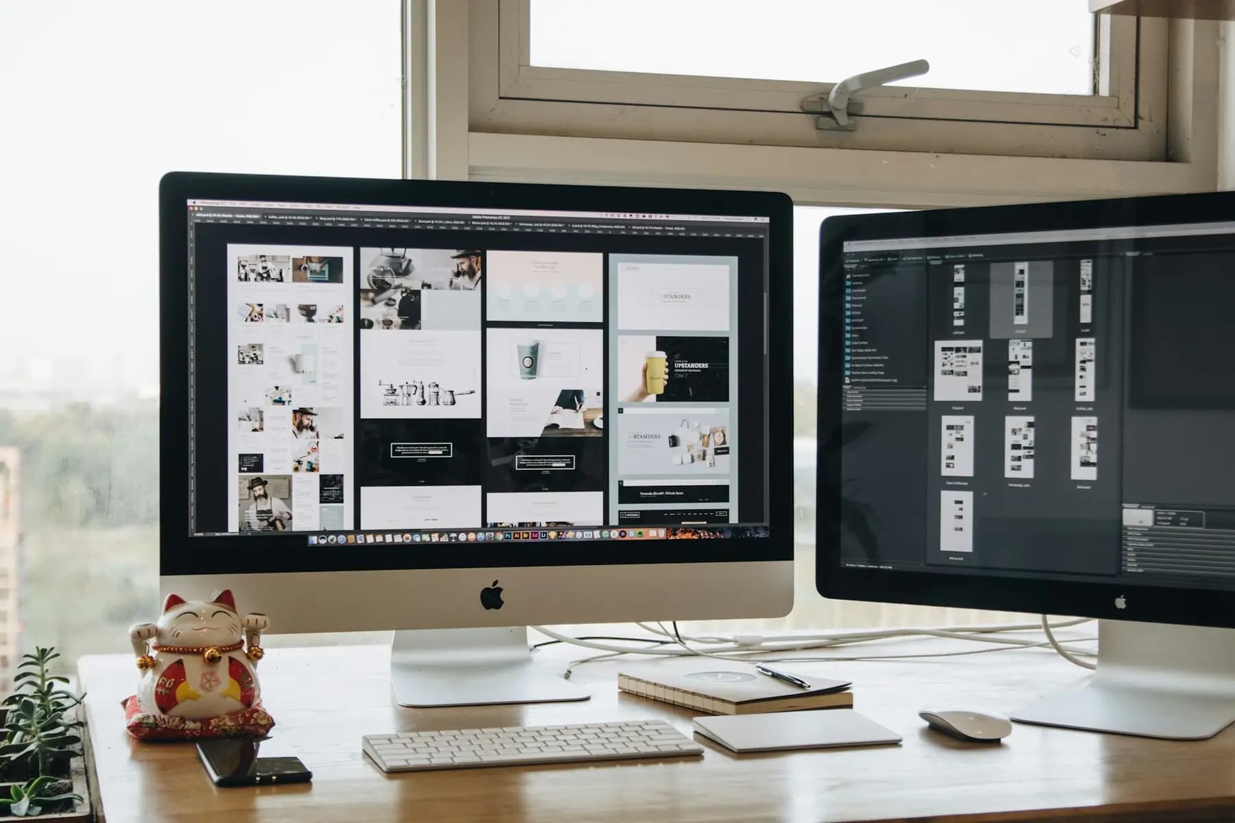 Two large monitors showing website layouts on a bright desk by a window